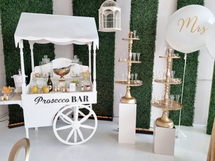 Elegancki kącik Prosecco Bar na przyjęciu, z białym wózkiem na kółkach, udekorowanym złotymi akcentami i szklanymi naczyniami. Na wózku znajdują się butelki prosecco, kieliszki, karafki z sokami oraz duża srebrna misa z lodem. Obok ustawiono złote stojaki na kieliszki, a w tle ściana z zielonego mchu dodaje stylowego charakteru. Nad wózkiem wisi ozdobna biała klatka, a po prawej stronie duży biały balon z napisem „Mrs”, co sugeruje, że jest to część dekoracji ślubnej lub wieczoru panieńskiego. Całość tworzy luksusową i nowoczesną aranżację na eleganckie przyjęcie