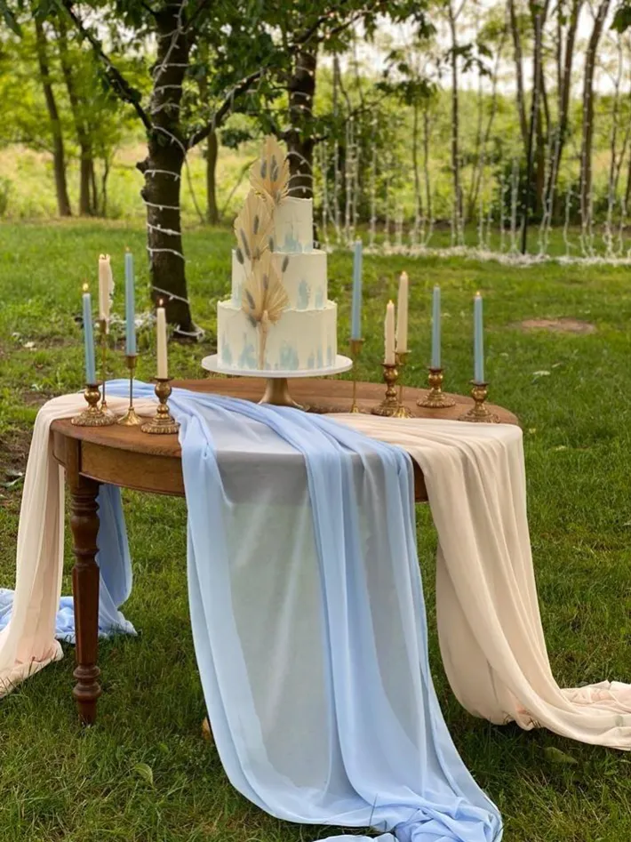 Rustykalny stół na świeżym powietrzu, udekorowany pastelowymi tkaninami w odcieniach beżu i błękitu, które swobodnie opadają na trawę. Na środku stoi elegancki, piętrowy tort weselny ozdobiony złotymi liśćmi palmowymi i błękitnymi detalami. Wokół ustawiono złote świeczniki z długimi, jasnoniebieskimi i kremowymi świecami. W tle widać drzewa oplecione białymi światełkami, tworząc romantyczną atmosferę w stylu boho